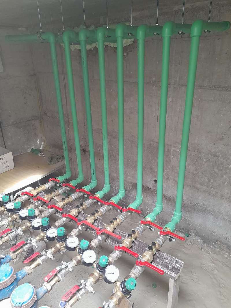 Zelene vertikale za vodu spojene na niz vodomjera s crvenim ventilima uz betonski zid.